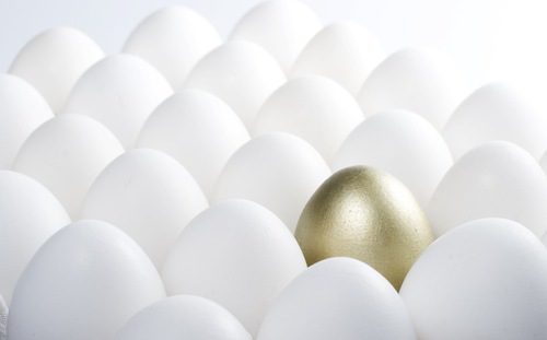 A gold egg is in the middle of many white eggs.